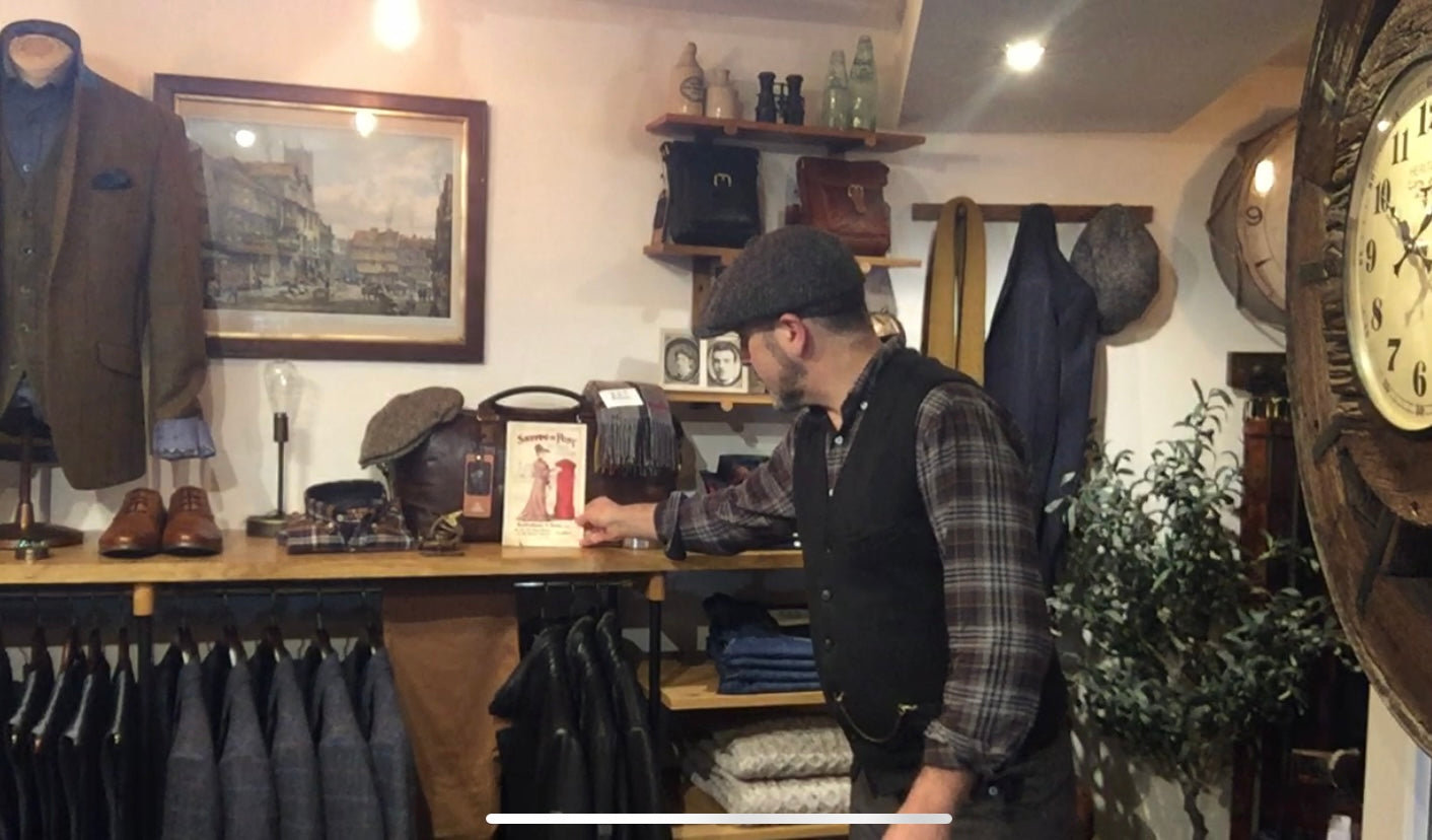The menswear corner inside Bodenhams of Ludlow, a tweed jacket on a stand, leather satchels and flat caps on a shelf, a member of staff in a flat cap and waistcoat